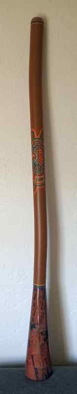 Photo of a didgeridoo stood up on top of an ottoman against a white wall. It is about 4 feet high and there is a lizard design made mostly of little dots of color. The wood is a medium brown.