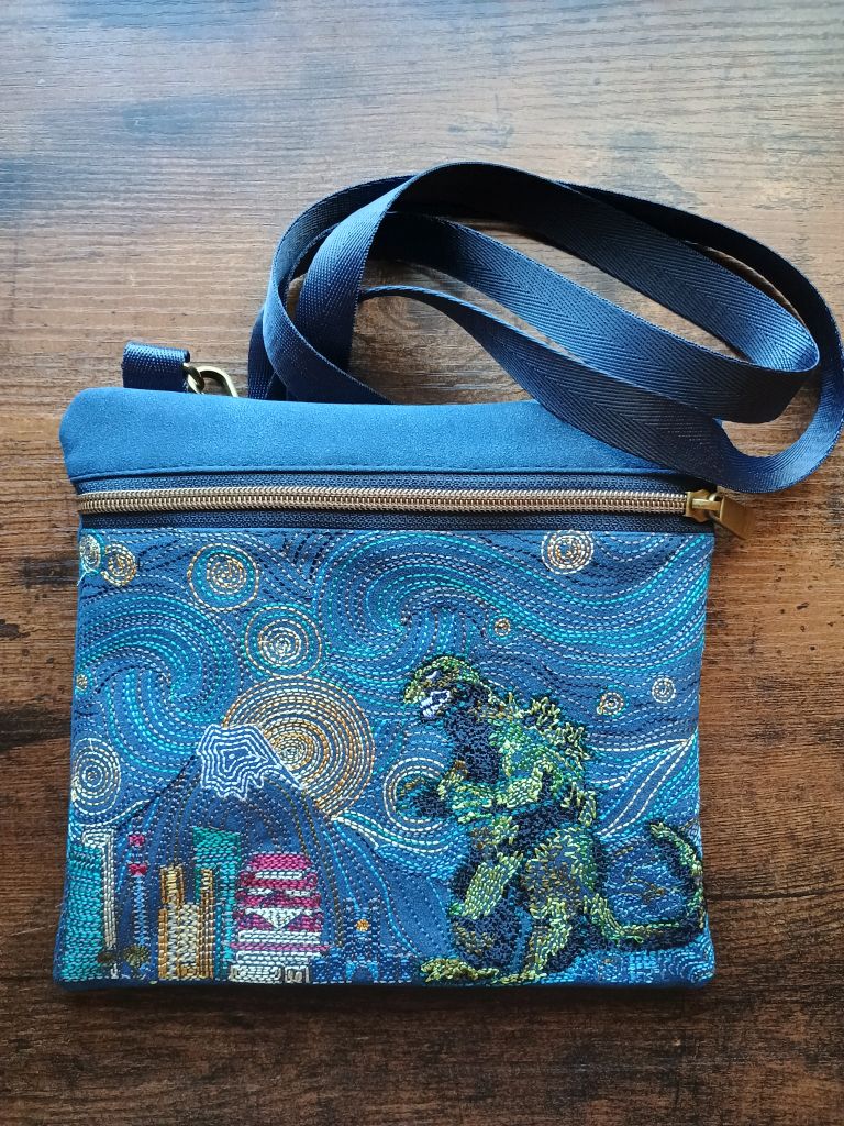 A dark blue purse with a zipper on the top side, long blue straps which are doubled up, and a scene featuring a sky reminiscent of Van Gogh's Starry Night painting but with Mt. Fuji and a city scene full of tall buildings on the left and an image of Godzilla on the right, heading towards the city. The whole scene is embroidered in threads of various colors.