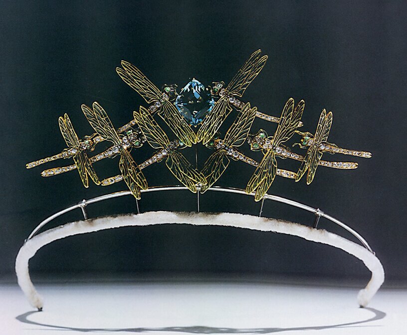 Tiara made by René Lalique featuring 4 dragonflies on each side of a diamond-shaped aquamarine. The dragonflies are flying at an upwards angle and they are decorated in diamonds, emeralds and their wings are enameled. The tiara is on a wire frame of silver metal and there is white velvet covering the wire where it would sit on the wearer's hair.