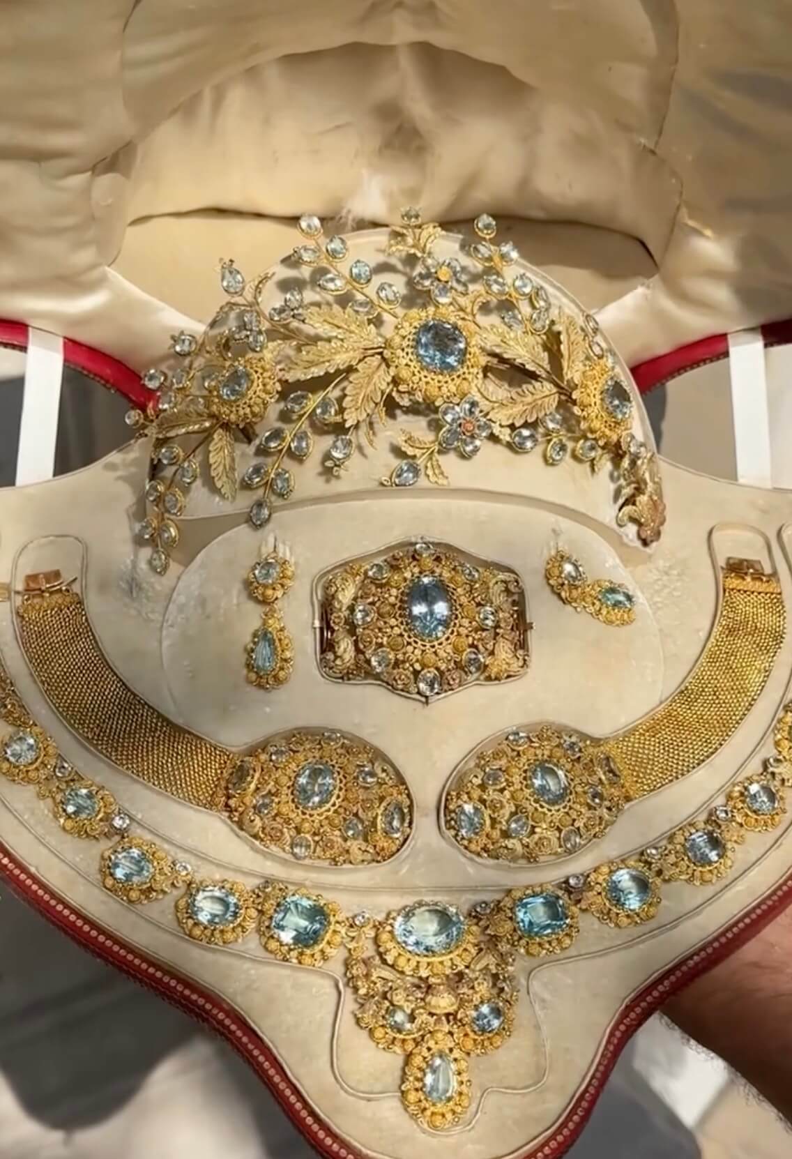 Gold and aquamarine parure in original case with tiara, necklace, brooch, earrings, 2 bracelets. The tiara design is sprays of aquamarine flowers and leaves, golden leaves, large round aquamarines in ornate circular frames. The earrings are studs and drops, each with aquamarines. The bracelets are gold mesh with large oval medallion clasps. The brooch is large and oval, set with an oval aquamarine. The necklace is ornate goldwork with oval aquamarines and a pendant in the middle that has 3 other aquamarines, set in ornate goldwork.