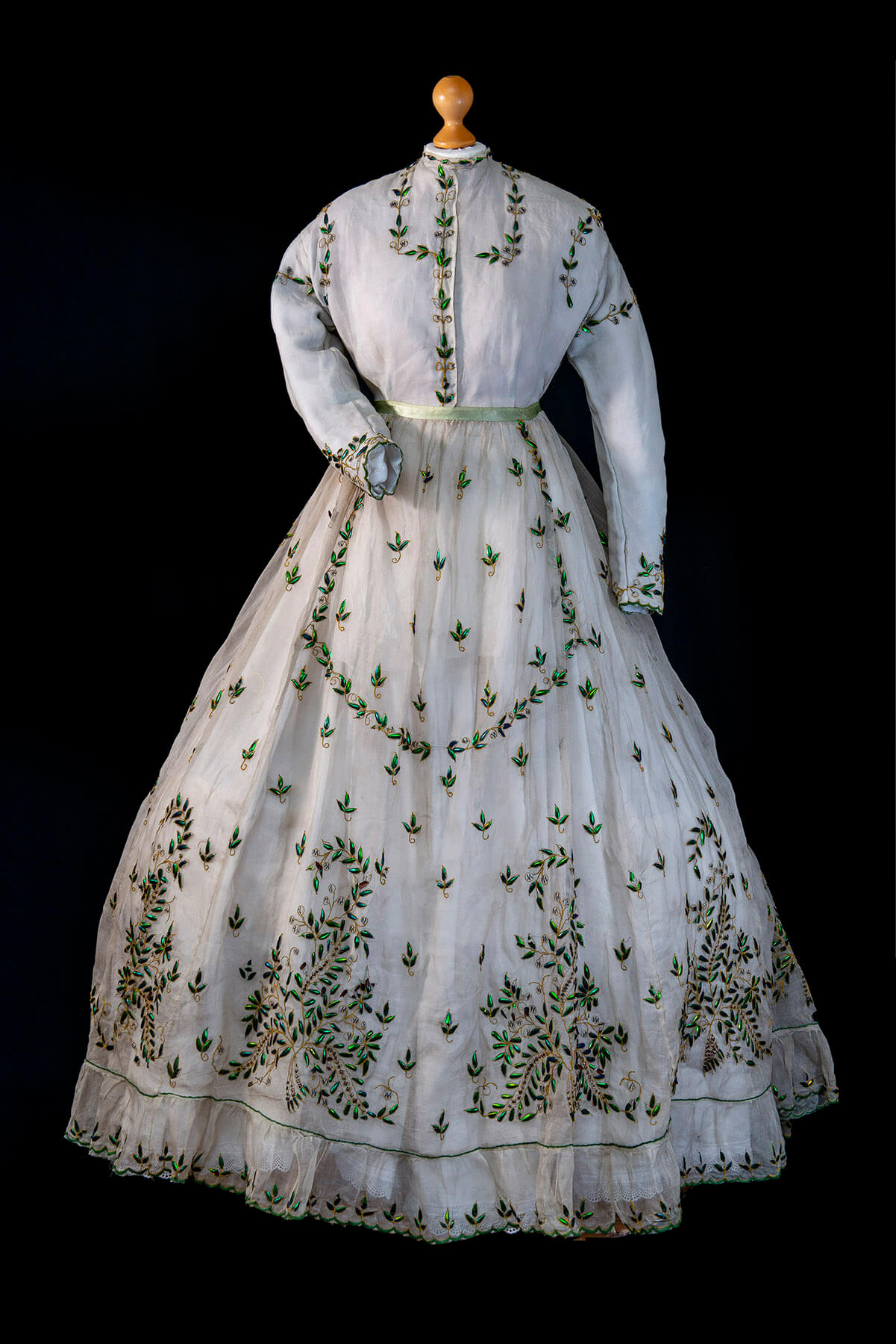 white dress from the 1800s decorated with iridescent beetles. There is a line of beetles down the front of the bodice, outlining the cuffs, neckline, shoulder areas. The skirt of the dress is covered in large and small motifs, some looking like foliage, all created from beetles.