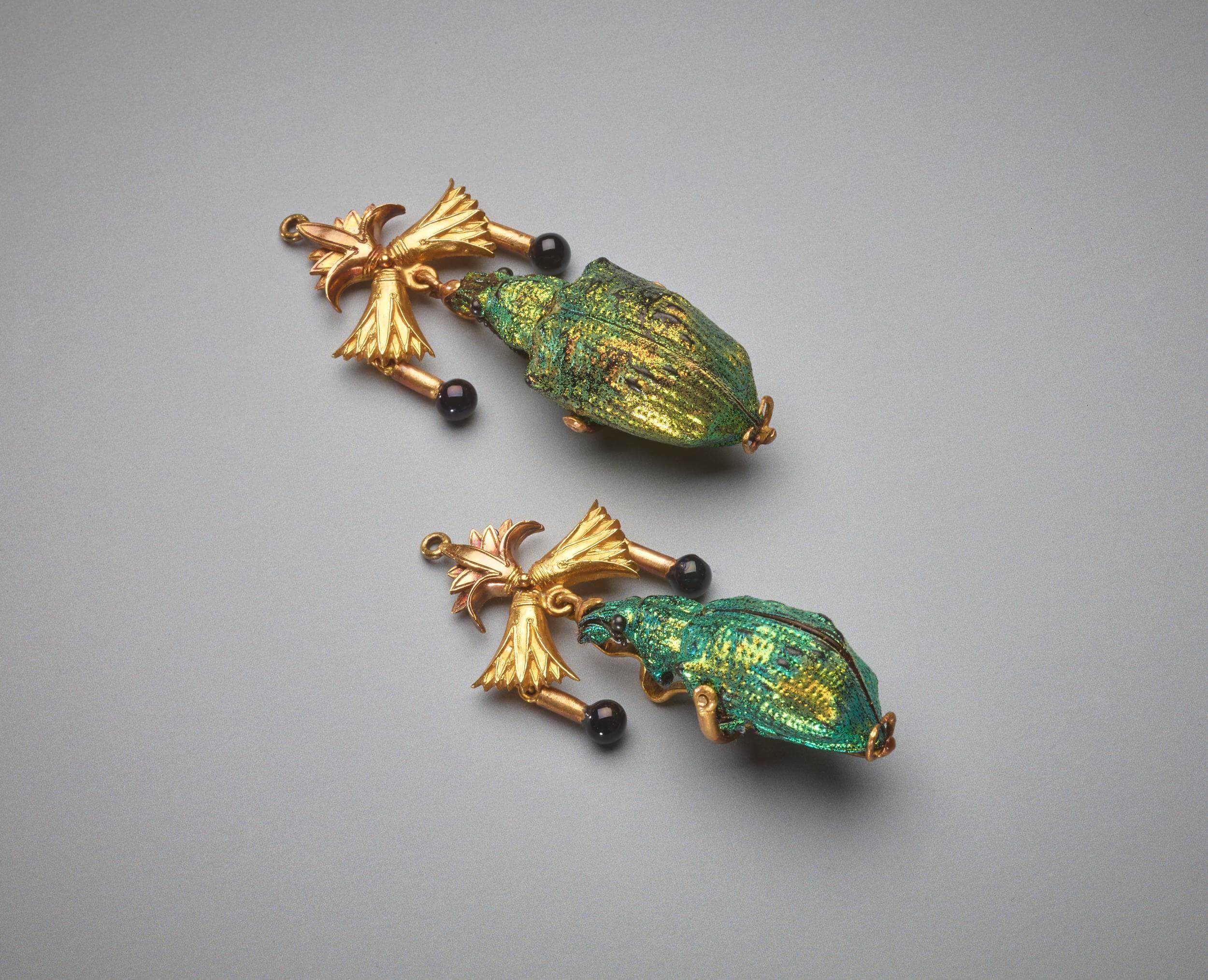 Earrings made from weevil shells. They are suspended from a fitting with one lotus blossom at top, two underneath with one facing left and one facing right and to each side of the weevil shell is a small gold rod with a black enamel bead at the bottom.