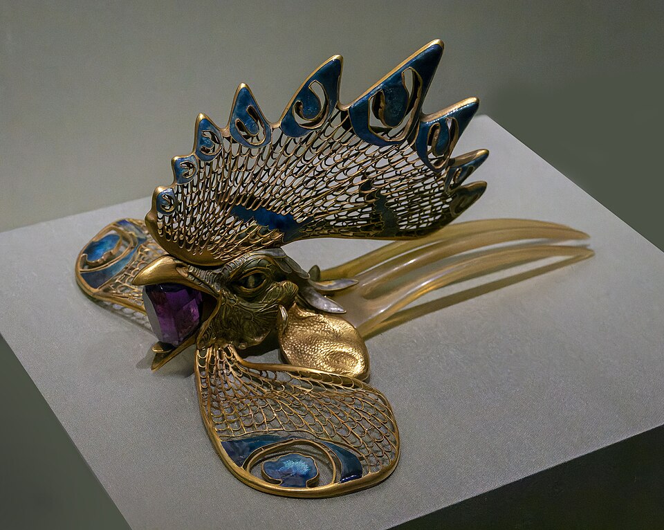 Side view of the Cockerel Diadem, a hair ornament featuring a three-pronged tan-colored hair comb with a golden rooster head attached. The rooster's comb is lacy goldwork with green, blue and enamel details. The wattles are lying flat and to the sides, also made of lacy goldwork and enamel detail. The rooster is holding a huge, faceted amethyst stone in its beak.