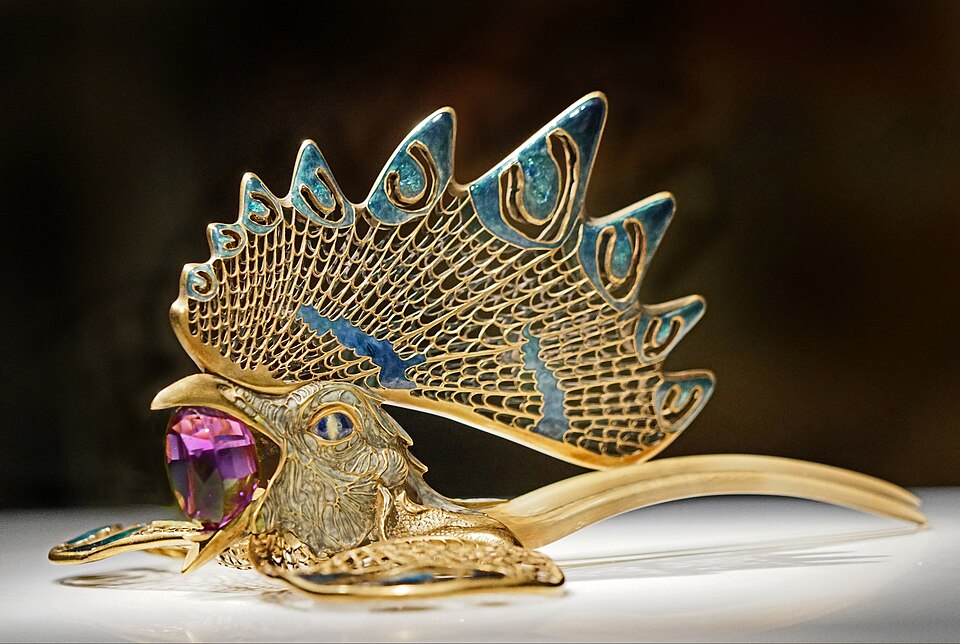 Side view of the Cockerel Diadem, a hair ornament featuring a three-pronged tan-colored hair comb with a golden rooster head attached. The rooster's comb is lacy goldwork with green and blue enamel details. The wattles are lying flat and to the sides, also made of lacy goldwork and enamel detail. The rooster is holding a huge, faceted amethyst stone in its beak.