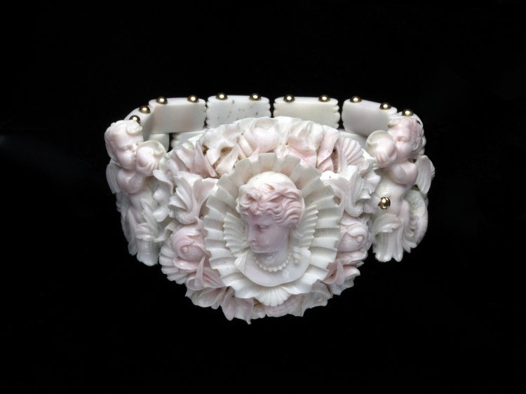 Close up view of the conch shell bracelet. The center piece is a woman's bust, head looking to the left. She has a pearl necklace, stiff lace ruff behind her, and her hair is in an updo. She is framed in a scallop shell and behind the scallop shell are other marine motifs like shells. To either side are cupids with seahorse tails, blowing conch shells.