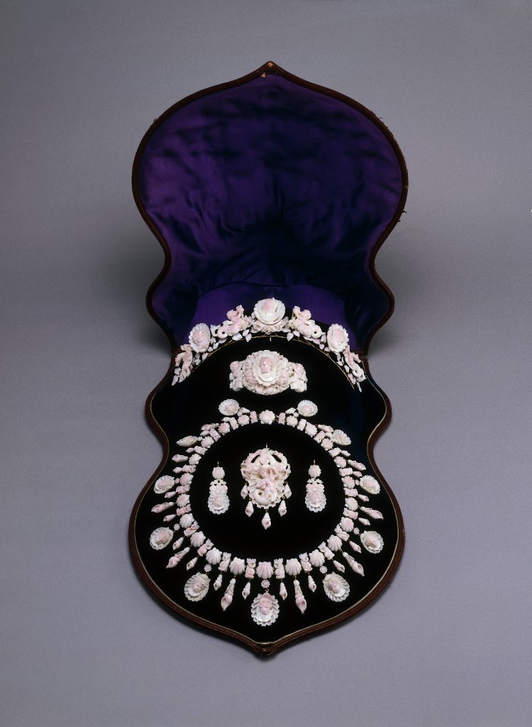 A picture of the entire parure in its blue velvet lined case. The parure consists of a tiara, bracelet, necklace, brooch (which converts to a pendant) and earrings, all carved out of conch shell.
