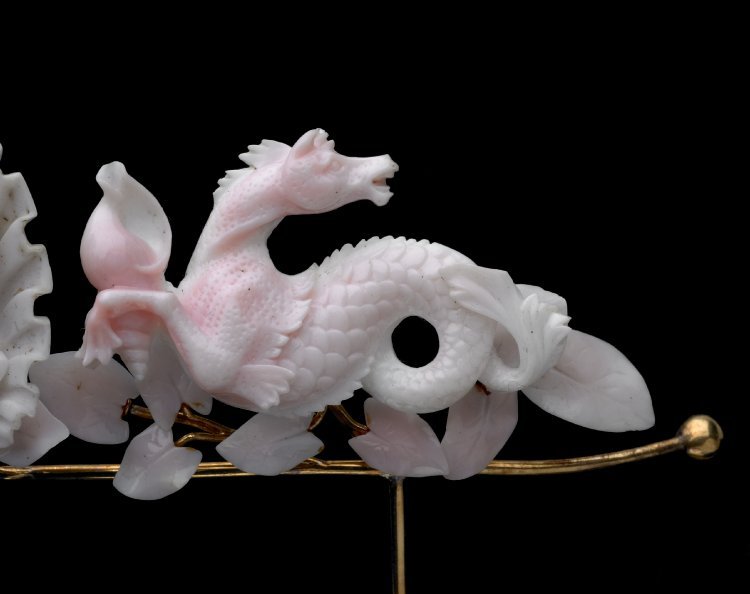 Close up of one of the seahorses on the tiara. It's head is turned to the right, looking over its back. It's front arm is in front of a conch shell that it appears to be holding. There are small leaves under the seahorse.