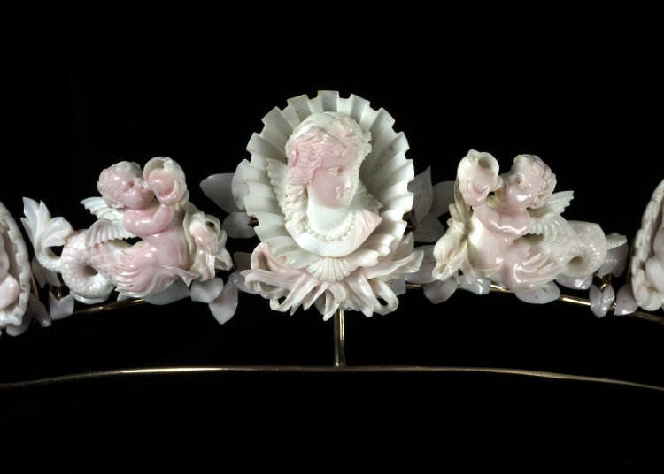 Close up image of a tiara made from conch shell. In the center is a woman's bust looking down and to the right. She is wearing a pearl necklace, has long hair which is pulled back from her face and you can see the sleeves of her dress on her shoulders. The bust is framed within a scallop shell. To either side of her are little cherubs with seahorse tails, blowing conch shells.