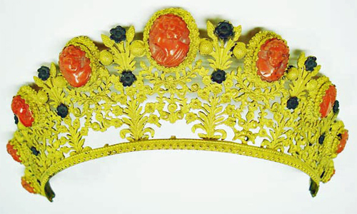 Image of an ornate tiara made of gilt bronze, coral cameos and black flowers. There are floral elements with stems and leaves with small black flowers. In between the floral elements are 9 coral cameos spaced around the tiara with various carved faces. The bronze is an obnoxious yellow color and with the red cameos and black flowers the tiara resembles the old Windows 3.1 Hot Dog Stand color scheme.