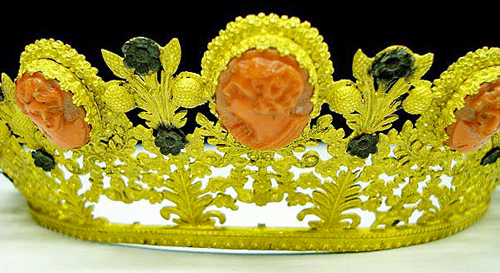 Image of an ornate tiara made of gilt bronze, coral cameos and black flowers. There are floral elements with stems and leaves with small black flowers. In between the floral elements are 9 coral cameos spaced around the tiara with various carved faces. The bronze is an obnoxious yellow color and with the red cameos and black flowers the tiara resembles the old Windows 3.1 Hot Dog Stand color scheme.