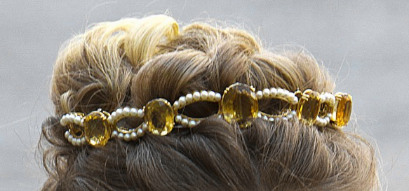 Tiara perched in blonde updo hairstyle. Gold tiara with large oval orange gemstones, either citrine or topaz, interspersed with seed pearl dividers. Each divider is made of a piece arching up and a piece arching down.
