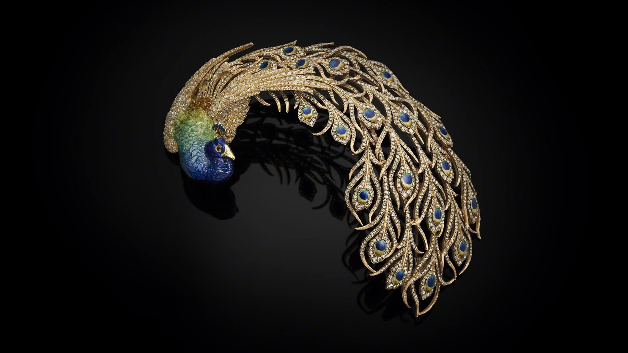 Decorative peacock headpiece made from gold, diamonds and enamel. The peacock's tail is curved to the side. The feathers are studded with diamonds but the eyes of the feathers are enameled in blue, yellow and gold. The body of the peacock is also enameled in shades of green and blue. The beak is golden.