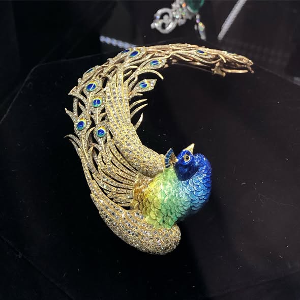 Decorative peacock headpiece made from gold, diamonds and enamel. The peacock's tail is curved to the side. The feathers are studded with diamonds but the eyes of the feathers are enameled in blue, yellow and gold. The body of the peacock is also enameled in shades of green and blue. The beak is golden.