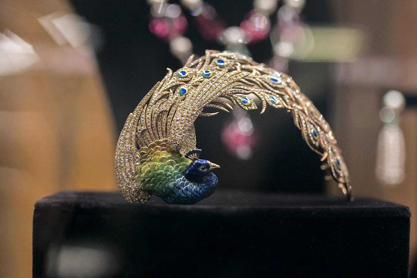 Decorative peacock headpiece made from gold, diamonds and enamel. The peacock's tail is curved to the side. The feathers are studded with diamonds but the eyes of the feathers are enameled in blue, yellow and gold. The body of the peacock is also enameled in shades of green and blue. The beak is golden.