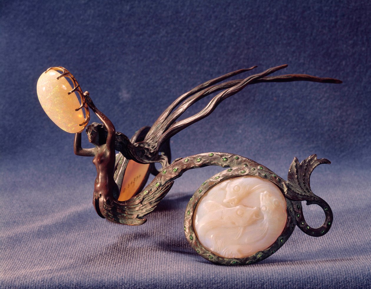 Bronze diadem in the shape of a siren. Her arms are raised up and holding a large oval opal above her head. She is naked to the waist and then her tail splits in two, each half forming one half of the diadem. The tails are embossed with shapes suggesting scales and are studded with small emeralds. They curve around round white opalescent pieces representing bubbles that are carved with multiple fish. The siren's hair flows out backward, forming 3 prongs which are used to fasten the diadem into the wearer's hair.
