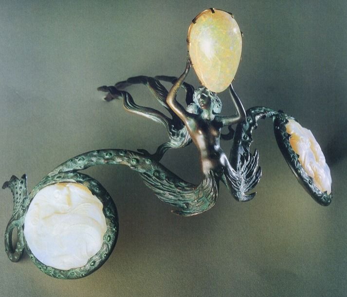 Bronze diadem in the shape of a siren. Her arms are raised up and holding a large oval opal above her head. She is naked to the waist and then her tail splits in two, each half forming one half of the diadem. The tails are embossed with shapes suggesting scales and are studded with small emeralds. They curve around round white opalescent pieces representing bubbles that are carved with multiple fish. The siren's hair flows out backward, forming 3 prongs which are used to fasten the diadem into the wearer's hair.