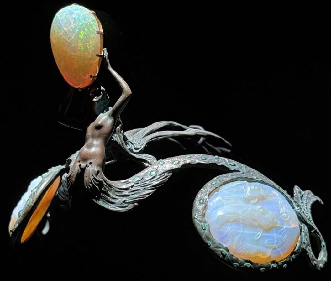 Bronze diadem in the shape of a siren. Her arms are raised up and holding a large oval opal above her head. She is naked to the waist and then her tail splits in two, each half forming one half of the diadem. The tails are embossed with shapes suggesting scales and are studded with small emeralds. They curve around round white opalescent pieces representing bubbles that are carved with multiple fish. The siren's hair flows out backward, forming 3 prongs which are used to fasten the diadem into the wearer's hair.