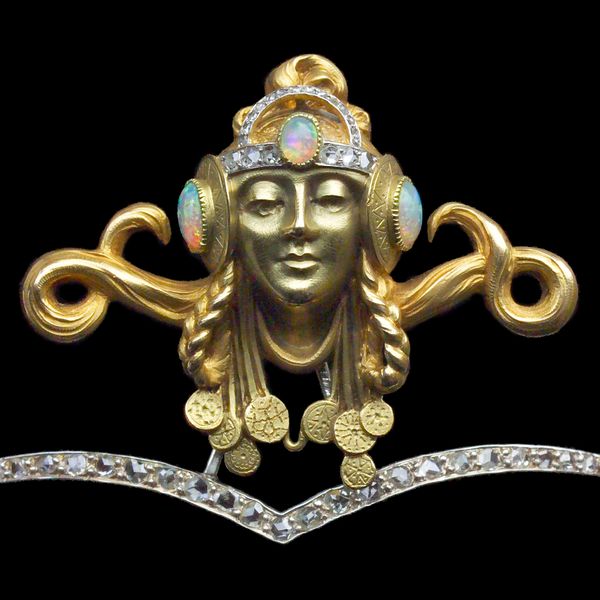 Close up image of the central portion of a tiara. The band is studded with diamonds and has 2 softly rounded arches coming down to a point in the center. It features the golden head of a woman which is finely detailed. She has an elaborate hairstyle with hair piled on top in an updo as well as braids hanging down and doubled up. Behind her head on either side appear to be long, thick, hanks of hair that are looped in circles at the lower part with the end of the hank pointing up. She appears to be wearing a necklace and there are several ribbons with medallions on each end around her neck. The medallions, which are all round, have designs on them like stars, lines, and dots. The figure is wearing a headpiece. Across her forehead is a diamond band with a large oval opal in the center. There is a smaller semi-circular diamond band rising up from this main band. On the sides, covering where her ears would be, there are large circular golden medallions engraved with angular line decorations with large round opals in the center.  