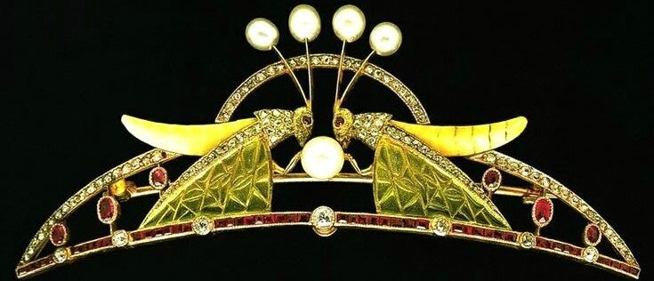 Grasshopper tiara in Art Nouveau style. 2 grasshoppers are facing each other in the center, with front legs resting on a large pearl. Their antennae are topped with pearls. The bodies are made of a yellow-green stone or enamel and the wings are green translucent enamel. Their eyes are rubies and the front of their bodies and the edges of their wings have diamonds. The bottome band of the tiara is rectangular cut rubies interspersed with round diamond and there are oval rubies in the central part of the tiara, on either end of the grasshoppers. There is a semi-circle of diamonds behind the grasshoppers.