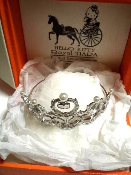 A view of the tiara from the top. It's placed in a case, resting on what looks like tissue paper. The outside of the case is orange. On the inside on a creamy satin is a printed image of Hello Kitty riding in a 2-wheeled contraption, pulled by a horse. She appears to be wearing the tiara. The tissue paper obscures part of the writing but you can see 