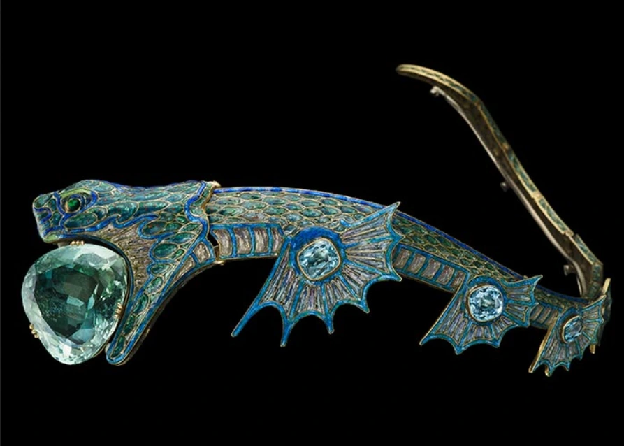 close up of sea dragon tiara. Gold diadem in the shape of a sea dragon with enamelled scales in pearl, green and blue tones, and a large triangular sea-green aquamarine being held in its jaws. The sea dragon is facing the left. You can see 3 sets of fins on the bottom, each one with a large blue aquamarine in the middle. It is articulated and the tail is pointing upwards.