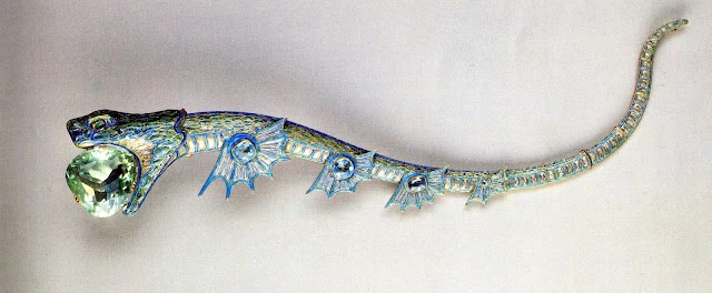 Sea dragon tiara fully stretched out and not on the tiara frame. Gold tiara in the shape of a sea dragon with enamelled scales in pearl, green and blue tones, and a large triangular sea-green aquamarine being held in its jaws. The sea dragon is facing the left. You can see 4 sets of fins on the bottom, each one with a blue aquamarine in the middle and getting progressively smaller as they go towards the tail.