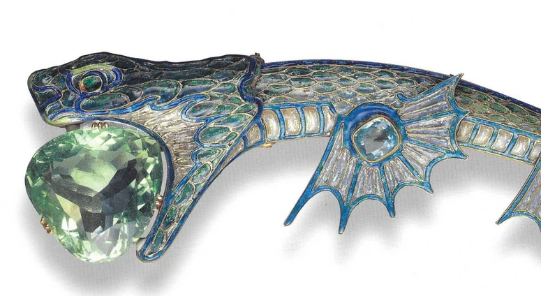 Close up of front portion of sea dragon tiara. Gold tiara in the shape of a sea dragon with enamelled scales in pearl, green and blue tones, and a large triangular sea-green aquamarine being held in its jaws. The sea dragon is facing the left. You can see 1 set of fins on the bottom right.