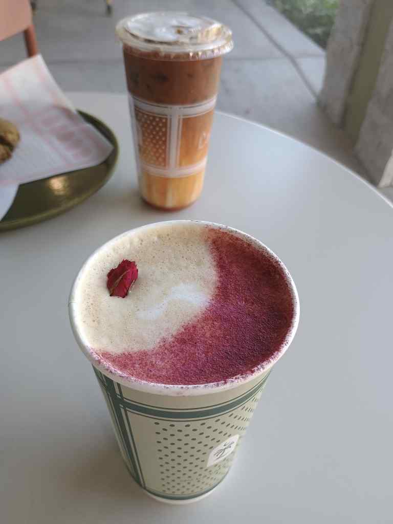 Photo looking down on a tall paper coffee cup with a green and white dots and bars design on a white table top. Inside the cup there is white froth on the left side as well as a tiny dried rosebud. On the right side there is dark rose-colored powder. In the background on the right, a cold coffee drink in a plastic cup can be seen. It is dark brown at the top and it gets gradually lighter towards the bottom. On the left there is a green plate with a waxed paper wrapper and you can just barely see part of a large, chunky cookie.