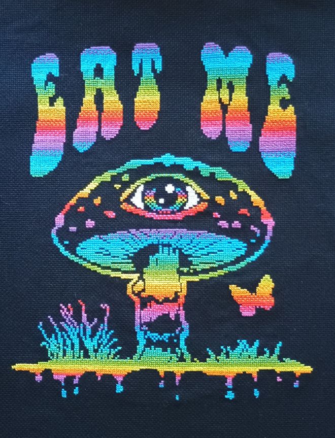 Cross stitch done on black 18 count Aida. Image is of a large mushroom with an eye in the middle. There are colored splotches on the mushroom top, the stalk has multicolored bands and the gills underneath are also multicolored. There is a small butterfly to the right. On the ground to either side of the mushroom are some grass-like plants, the ones on the left are taller. The ground appears to have drip marks falling towards the bottom. Above the mushroom are the words EAT ME in a 1970s kind of bubbly font. Everything is done in a bright rainbow gradient. It all looks very psychedelic.
