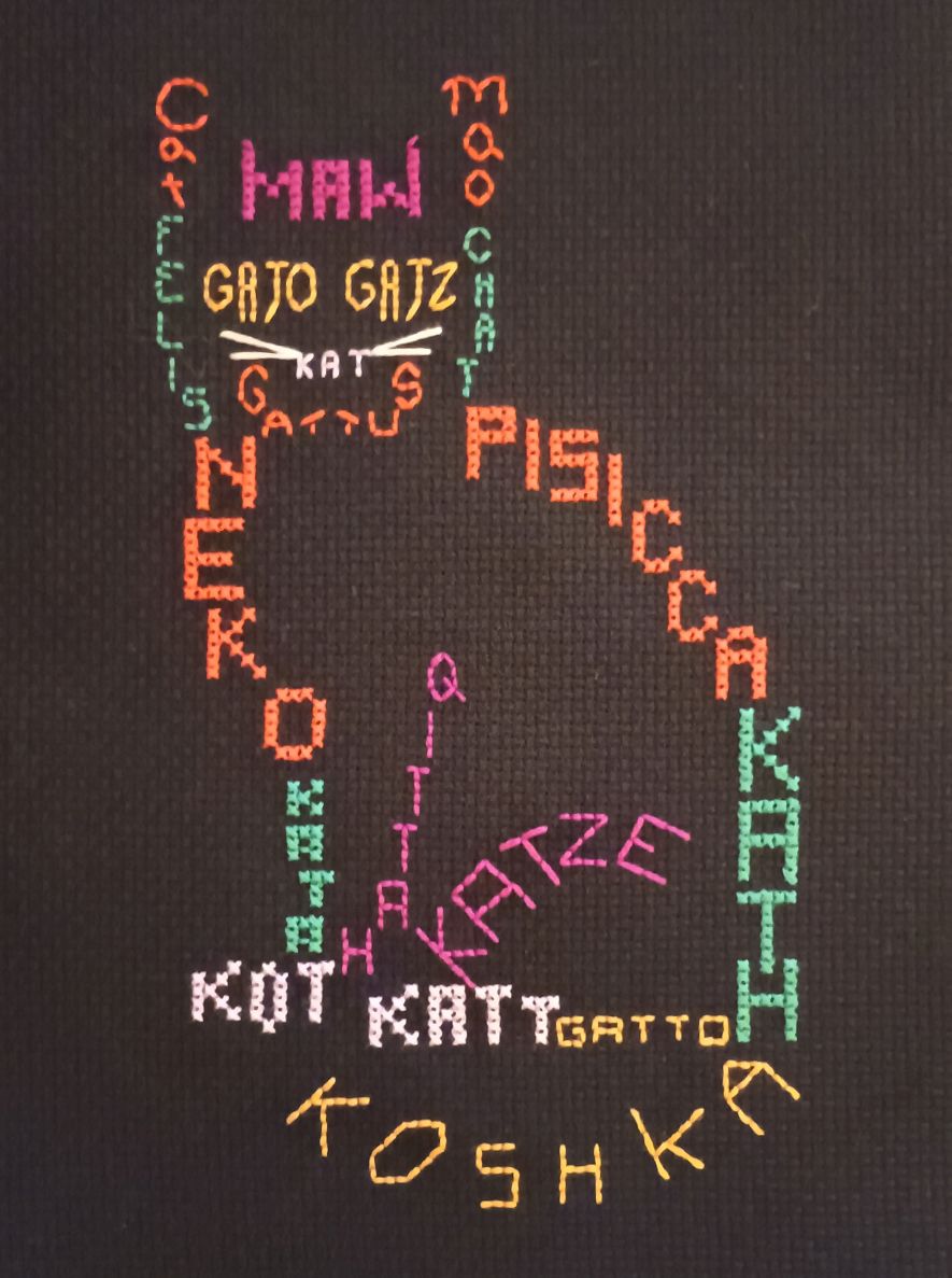 A cross-stitched outline of a cat. The cat is sitting up in profile but with the head facing the viewer. The outline is made out of the word for cat in different languages and colors. Here are the body parts, their color and the words and the language. Ears, orange-red: Cat (English) & Mao (Chinese);
Forehead, plum: Maw (Egyptian); Sides of face, aquamarine: Felis (Latin) & Chat (French); Eyes, topaz: Gato (Portuguese) & Gatz (Armenian); Nose, lavender: Kat (Dutch); Jaws, orange-red: Gattus (Yiddish); Body (top half), orange red: Neko (Japanese) & Pisicca (Romanian); Body (bottom half), aquamarine: Kata (Greek) & Kath (Welsh); Legs, plum: Qittah (Arabic) & Katze (German); Feet, lavender: Kot (Polish) & Katt (Norwegian); Bottom of body, topaz: Gatto (Italian); Tail, topaz: Koshka (Russian).