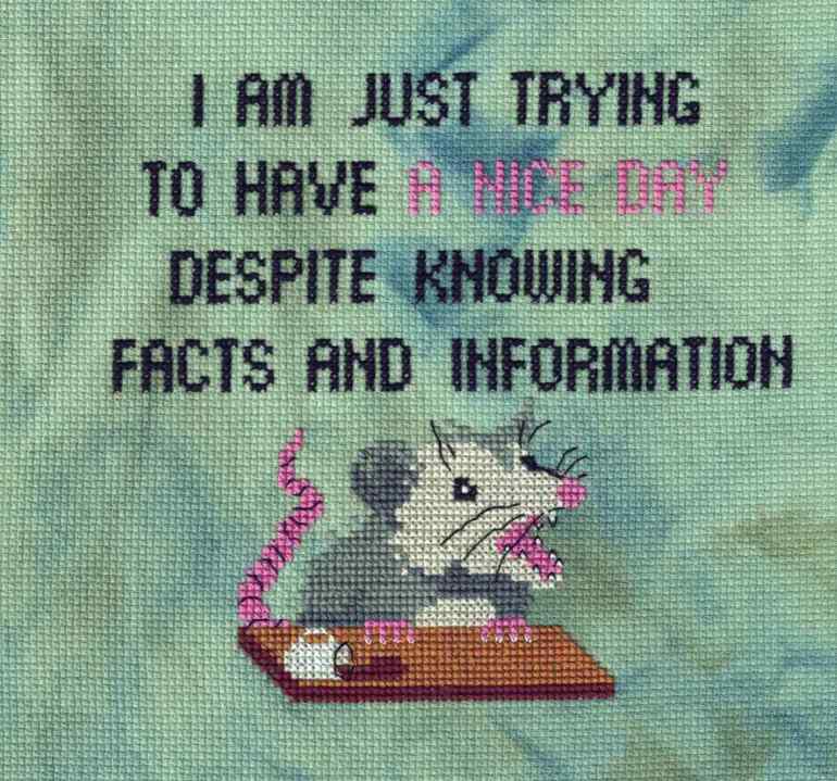 Cross stitch done on 14 count Aida. The color is Tempest, described as light aqua-green with blue and yellow-green splashes. At the top in block capital letters: I AM JUST TRYING TO HAVE A NICE DAY DESPITE KNOWING FACTS AND INFORMATION. The letters are in black except for A NICE DAY which is in pink. Under that is a possum sitting at a desk top. You can see his upper half, his hands are gripping the desk and his tail is visible on the left. His mouth is open and he's looking slightly off to the right. There is a white coffee cup on the desk which has tipped over and spilled a dark brown liquid.