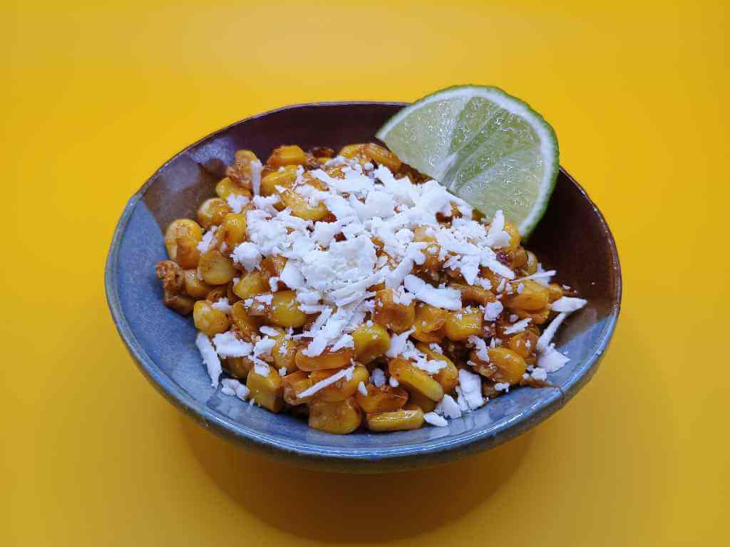 Photograph of a small shallow bowl with elote, a Mexican corn dish. The bowl is glazed in shades of blue and brown. The elote is yellow corn kernels that have been roasted until slightly brown. There is some red coloring from the spices. There is white cheese sprinkled on top. A lime wedge sits on top of the elote on the upper right. The background is a bright, deep yellow.